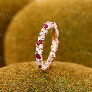 May include: A rose gold ring with alternating oval-cut red and round white gemstones. The ring is set on a textured, golden-brown surface. The gemstones are evenly spaced around the band.