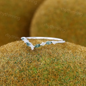 K&ouml;nnte beinhalten: Ein zarter Silberring mit einem geschwungenen Design, das eine Reihe von gr&uuml;nen Steinen in Marquise-Form und kleinere runde wei&szlig;e Steine aufweist.