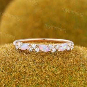 Peut inclure: Une bague en or rose ornée d'une rangée d'opales ovales et de petits diamants ronds. Les opales présentent une base blanc laiteux avec des éclats roses et bleus. La bague est posée sur une surface texturée brun doré.