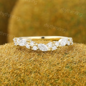 May include: A curved gold band ring featuring a cluster of clear, sparkling marquise-cut and round-cut stones arranged in a delicate, asymmetrical pattern along the top half of the band, creating a shimmering, multi-faceted appearance.
