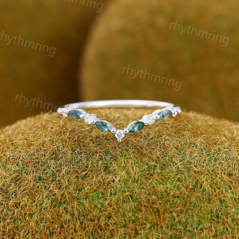 May include: A delicate silver ring with a v-shaped design. The ring is adorned with small, green marquise-shaped gemstones and a single, round diamond in the center.