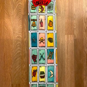 Puede incluir: Una tabla de monopatín con un diseño colorido de cartas de Loteria. La tabla muestra varias cartas ilustradas con texto en español, incluyendo "La Lotería" en la parte superior. La tabla está sobre una superficie de madera.