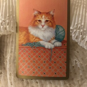 Op de afbeelding: Een close-up van een witte en oranje kat met groene ogen, liggend op een rood-wit gepatroneerd oppervlak. De kat houdt een bol wol in zijn poten.