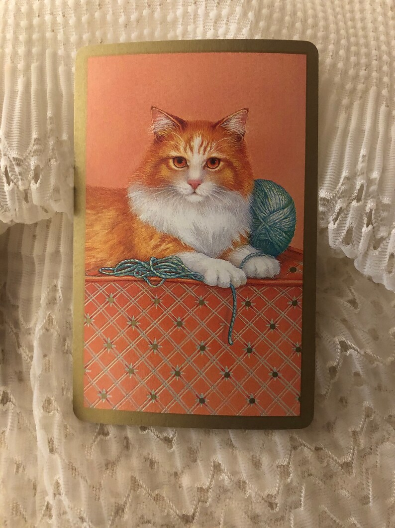Op de afbeelding: Een close-up van een witte en oranje kat met groene ogen, liggend op een rood-wit gepatroneerd oppervlak. De kat houdt een bol wol in zijn poten.