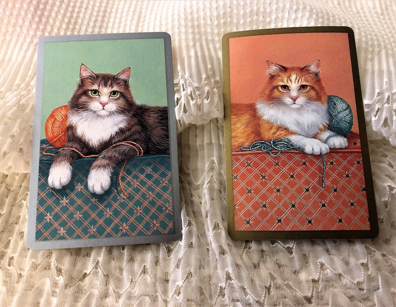 Op de afbeelding: Twee kaarten met afbeeldingen van katten die met garen spelen. De kat op de linker kaart is bruin en wit, en de kat op de rechter kaart is oranje en wit. Beide katten liggen op een gepatroneerd oppervlak.