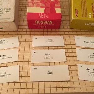 May include: A set of vocabulary flashcards for learning Russian, German, and Latin. The cards are white with black text and are arranged on a brown gridded surface. The cards feature words and their translations in the respective languages. The cards are from the Vis-Ed brand.