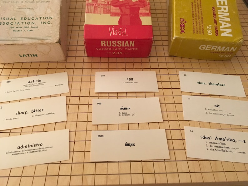 May include: A set of vocabulary flashcards for learning Russian, German, and Latin. The cards are white with black text and are arranged on a brown gridded surface. The cards feature words and their translations in the respective languages. The cards are from the Vis-Ed brand.