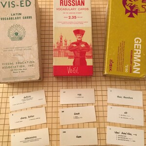 May include: Three boxes of vocabulary flashcards for learning Russian, German, and Latin. The boxes are red, yellow, and white with illustrations and text. The flashcards are visible on a brown grid surface.