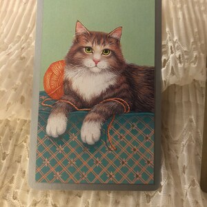 Op de afbeelding: Een bruin-witte tabbykat met groene ogen ligt op een groen-bruin gepatroneerd oppervlak. De kat speelt met een bolletje oranje garen.