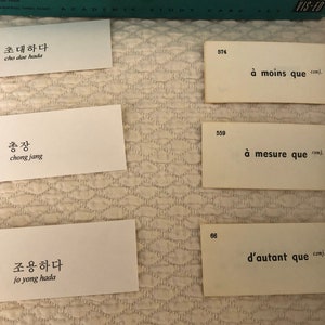 以下が含まれることがあります： 韓国語とフランス語の単語とフレーズが書かれたフラッシュカードのセット。カードは白地に黒文字。韓国語の単語はハングル、韓国のアルファベットで書かれています。フランス語の単語はラテン文字で書かれています。カードは821、822、746、574、559、66と番号が付けられています。