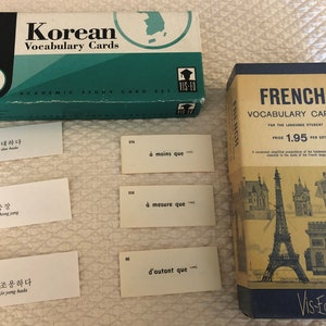 以下が含まれることがあります： 韓国語とフランス語の単語フラッシュカードが2箱あります。韓国語の箱は緑色で、韓国の地図が描かれています。フランス語の箱は黄色で、エッフェル塔と凱旋門の画像が描かれています。箱には「Korean Vocabulary Cards」と「French Vocabulary Cards」と書かれています。
