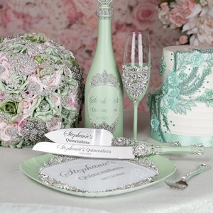 May include: A green and silver cake knife and server set with the text "Stephanie's Quinceañera 04/12/2024" on the handles. The set is on a green plate with a silver trim. A green bottle with a silver crown and the text "Stephanie's Quinceañera 04/12" is in the background.