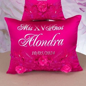 May include: Two fuchsia satin pillows, one stacked on the other. Each pillow features the text "Mis XV Años Alondra 10/05/2024" in silver script. Floral embellishments with pink roses and beaded accents adorn the lower portion of each pillow.