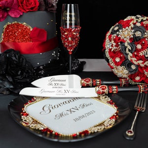 May include: A black and red cake with a gold crown on top. A red and gold bouquet of flowers, a silver cake knife, a silver cake server, and a red and gold wine glass are on a black surface. The cake knife, cake server, and wine glass are engraved with the text "Giovanna Mis XV Años 10/08/2023".