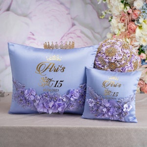 May include: Two light purple satin pillows featuring gold text that reads Ari's Sweet 15 and 10/28/2023. Each pillow is decorated with a curved arrangement of light purple fabric flowers, lace, and beads. A small gold crown sits behind the larger pillow.