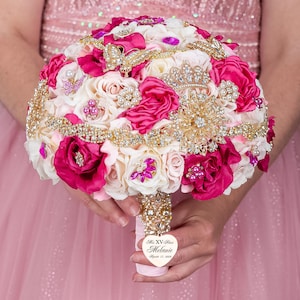 May include: A pink and white bridal bouquet with gold accents. The bouquet is decorated with rhinestones, pearls, and brooches. A heart-shaped tag with the text "Miss XV Kiss Melanie August 17, 2019" is attached to the bouquet.