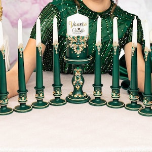 May include: A set of 10 green candle holders with gold accents and white candles. The candles are decorated with gold and green embellishments. The center candle has a white label that reads "Vanessa Quince".