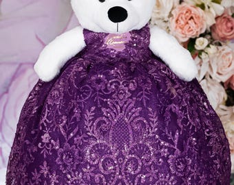 Könnte beinhalten: Ein weißer Plüsch-Teddybär mit einem goldfarbenen Diadem und einem dunkelvioletten Kleid. Das Kleid ist mit einem komplizierten floralen Spitzenmuster und glitzernden Pailletten versehen. Der Bär hat schwarze Augen und eine schwarze Nase.