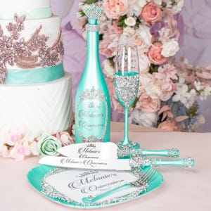 May include: A turquoise and silver cake knife set with a crown design and the text "Melanie's Quinceanera". The set includes a cake knife, a champagne bottle, and a champagne flute.