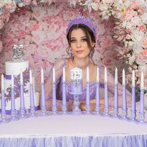 May include: A quincea&ntilde;era celebration table setting with a purple tablecloth, a white cake with a silver "15" on top, and a row of purple candle holders with white candles. The centerpiece is a purple candle holder with a white candle that says "Our Very Special Ashley".