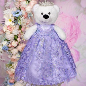 May include: A white teddy bear wearing a lavender dress with a sparkly, floral design and a silver crown. The dress has a lace overlay and the bear is standing in front of a pink and white floral background.