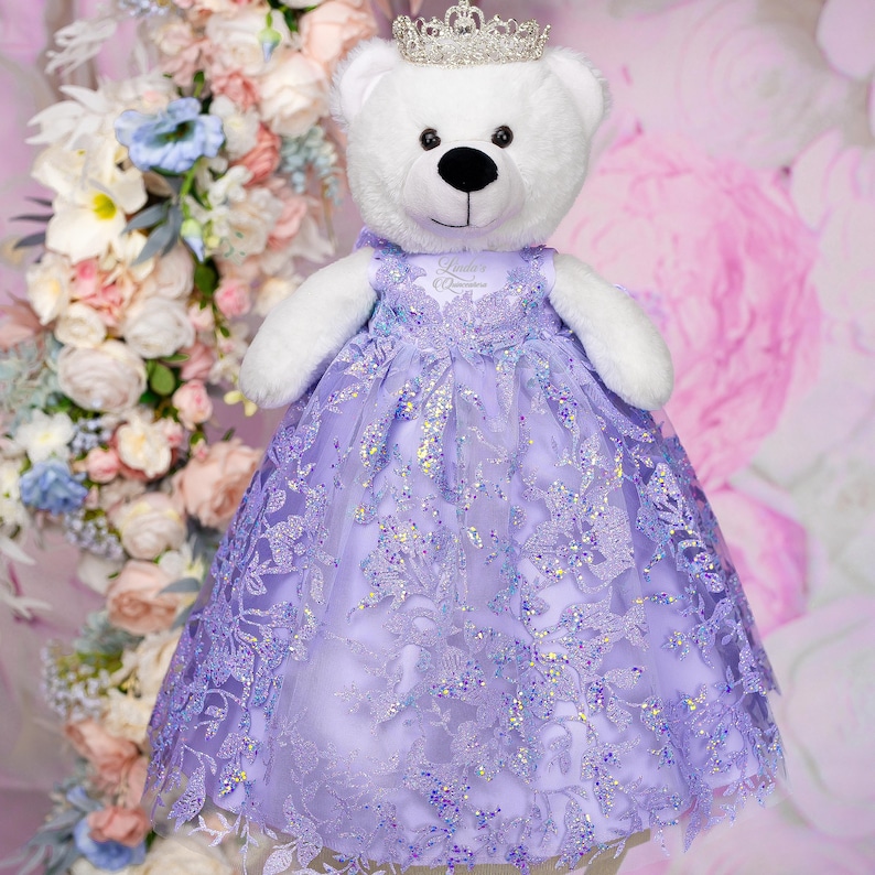 May include: A white teddy bear wearing a lavender dress with a sparkly, floral design and a silver crown. The dress has a lace overlay and the bear is standing in front of a pink and white floral background.