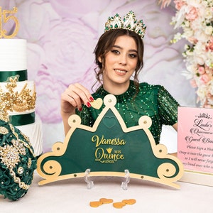 May include: A green and gold crown-shaped guest book with the text "Vanessa Quince MIS AÑOS" and a heart-shaped opening for guests to drop in their wishes. The crown is on a white table with a gold and green cake and a bouquet of flowers in the background.
