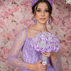 May include: A woman wearing a lavender gown with a sparkling bodice and a purple tiara holds a bouquet of lavender roses with rhinestones. The bouquet has a silver heart charm with the text "Mis XV A&ntilde;os Aubrey".