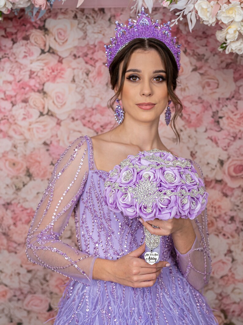 May include: A woman wearing a lavender gown with a sparkling bodice and a purple tiara holds a bouquet of lavender roses with rhinestones. The bouquet has a silver heart charm with the text "Mis XV A&ntilde;os Aubrey".