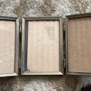 May include: A triptych photo frame featuring three rectangular panels connected by metal hinges. Each frame has a metallic gold-toned finish with a beaded edge detail and contains a plain, light brown cardboard insert behind clear glass.