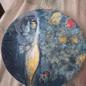 May include: Round decorative plate featuring a painting with a blue and yellow color scheme. The artwork depicts a figure with a long white garment, a rooster, and floral elements. The artist's signature is visible on the bottom left.
