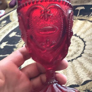 Könnte beinhalten: Ein leuchtend roter Glaskelch mit einem erhabenen, strukturierten Muster aus herzförmigen Motiven und Perlenverzierungen entlang des Randes und des Stiels. Das Gefäß hat einen breiten, runden Fuß und einen dicken, stabilen Stiel.