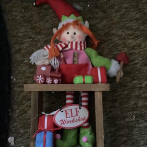 May include: A plush elf figurine with orange yarn hair, a red hat, and a striped scarf, sitting on a wooden workbench. The elf holds a red mallet and is surrounded by miniature wooden toys, gifts, and a sign that reads ELF Workshop.