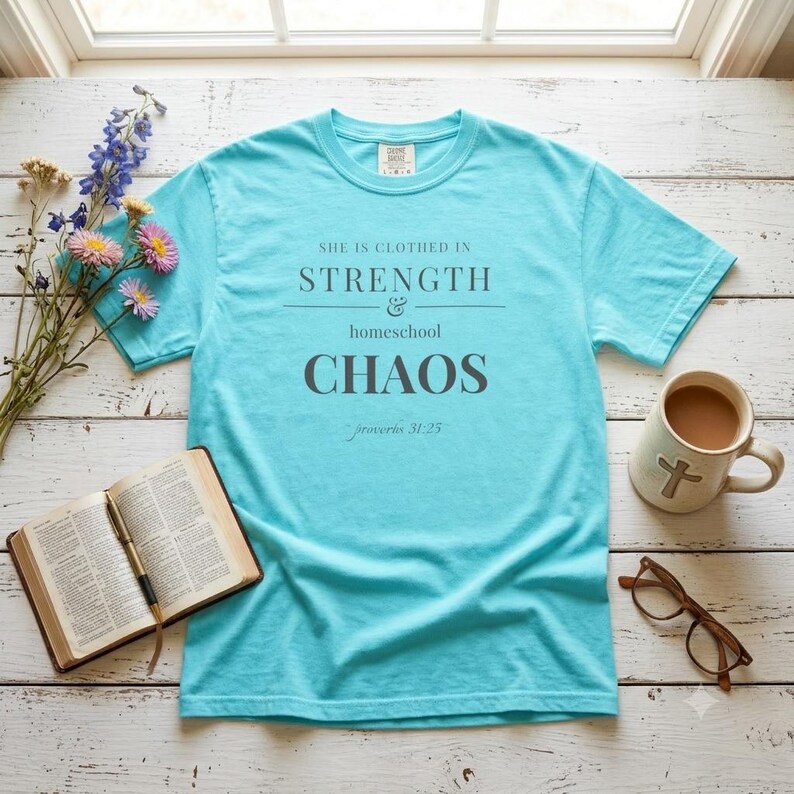 May include: A bright turquoise short-sleeve t-shirt featuring the dark gray text: SHE IS CLOTHED IN STRENGTH & homeschool CHAOS proverbs 31:25. The shirt has a crew neckline and a standard fit, displayed as a flat lay on a white wooden surface.