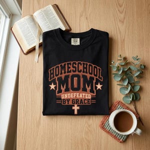 May include: A black crew neck t-shirt featuring the text "HOMESCHOOL MOM UNDEFEATED BY GRACE" in a bold, collegiate-style font. The design includes two stars on either side of the word "MOM" and a small cross symbol centered below the text.