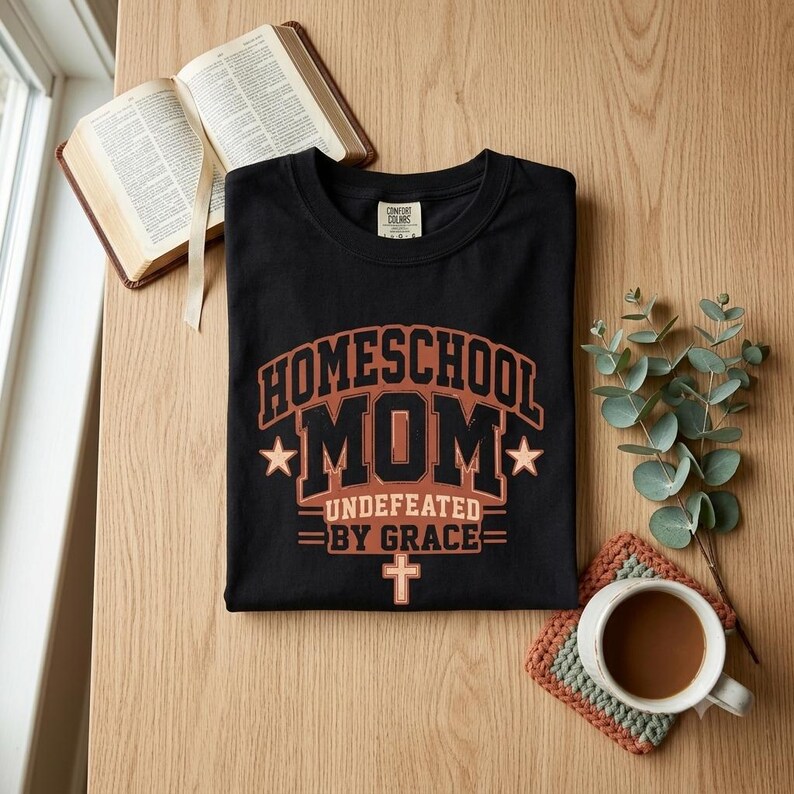 May include: A black crew neck t-shirt featuring the text "HOMESCHOOL MOM UNDEFEATED BY GRACE" in a bold, collegiate-style font. The design includes two stars on either side of the word "MOM" and a small cross symbol centered below the text.
