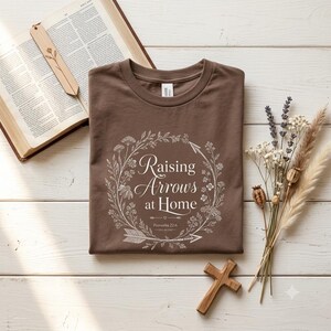 May include: A brown t-shirt featuring a white graphic of a floral wreath surrounding the text "Raising Arrows at Home" and "Proverbs 22:6". The shirt is displayed alongside an open book with a wooden bookmark and a small wooden cross.