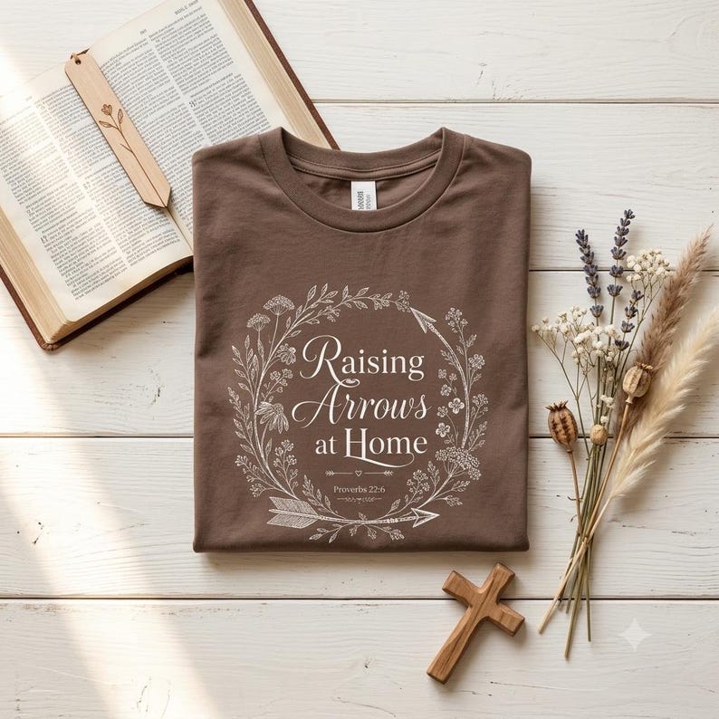 May include: A brown t-shirt featuring a white graphic of a floral wreath surrounding the text "Raising Arrows at Home" and "Proverbs 22:6". The shirt is displayed alongside an open book with a wooden bookmark and a small wooden cross.