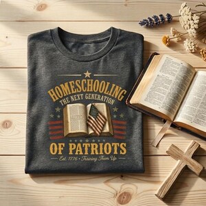 K&ouml;nnte beinhalten: Ein dunkelgraues T-Shirt mit einer Grafik, die ein offenes Buch, eine kleine amerikanische Flagge und den Text "HOMESCHOOLING THE NEXT GENERATION OF PATRIOTS Est. 1776 Training Them Up" zeigt. Das Design enth&auml;lt Sterne sowie rote und blaue Streifen.