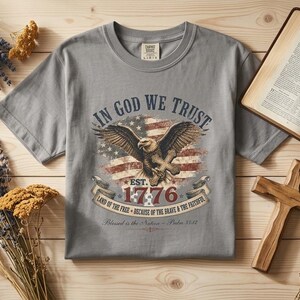 May include: A grey short-sleeved t-shirt featuring a graphic of an eagle, an American flag, and the text In God We Trust, Est. 1776, Land of the Free, Because of the Brave & the Faithful, and Blessed is the Nation - Psalm 33:12.