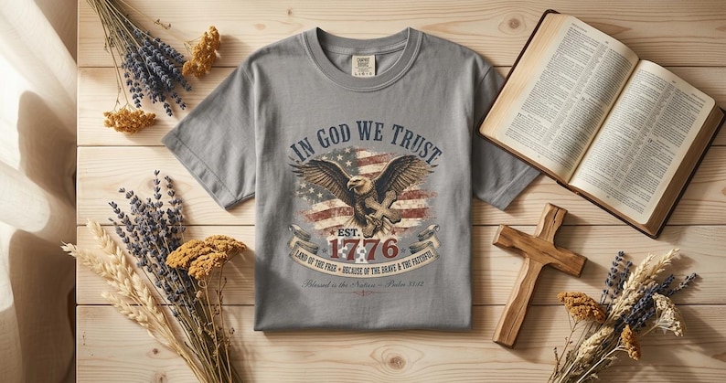 May include: A grey short-sleeved t-shirt featuring a graphic of an eagle, an American flag, and the text In God We Trust, Est. 1776, Land of the Free, Because of the Brave & the Faithful, and Blessed is the Nation - Psalm 33:12.