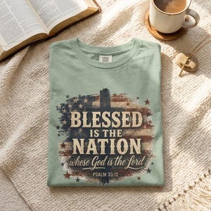 K&ouml;nnte beinhalten: Ein salbeigr&uuml;nes T-Shirt mit einer verwaschenen Grafik einer amerikanischen Flagge und einer Kreuz-Silhouette. Der Text lautet BLESSED IS THE NATION whose God is the Lord PSALM 33:12 in einer Mischung aus Serifenschrift und Schreibschrift mit Sternen.