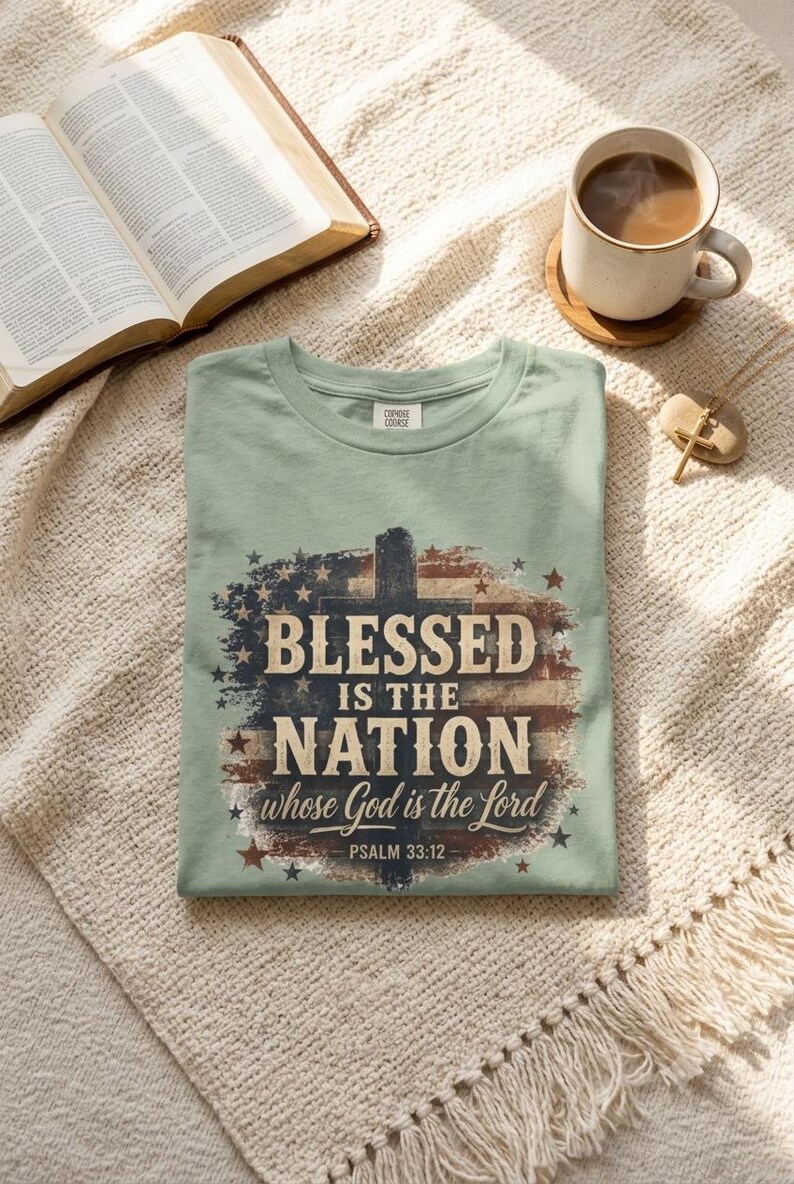 K&ouml;nnte beinhalten: Ein salbeigr&uuml;nes T-Shirt mit einer verwaschenen Grafik einer amerikanischen Flagge und einer Kreuz-Silhouette. Der Text lautet BLESSED IS THE NATION whose God is the Lord PSALM 33:12 in einer Mischung aus Serifenschrift und Schreibschrift mit Sternen.