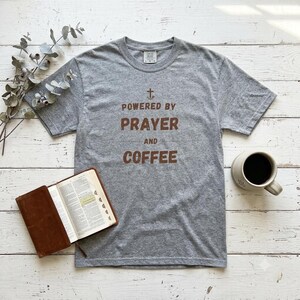 May include: A heather gray short-sleeve t-shirt featuring a brown graphic print that reads "POWERED BY PRAYER AND COFFEE" with a small cross symbol above the text. The shirt has a crew neckline and a standard fit, displayed on a flat surface.