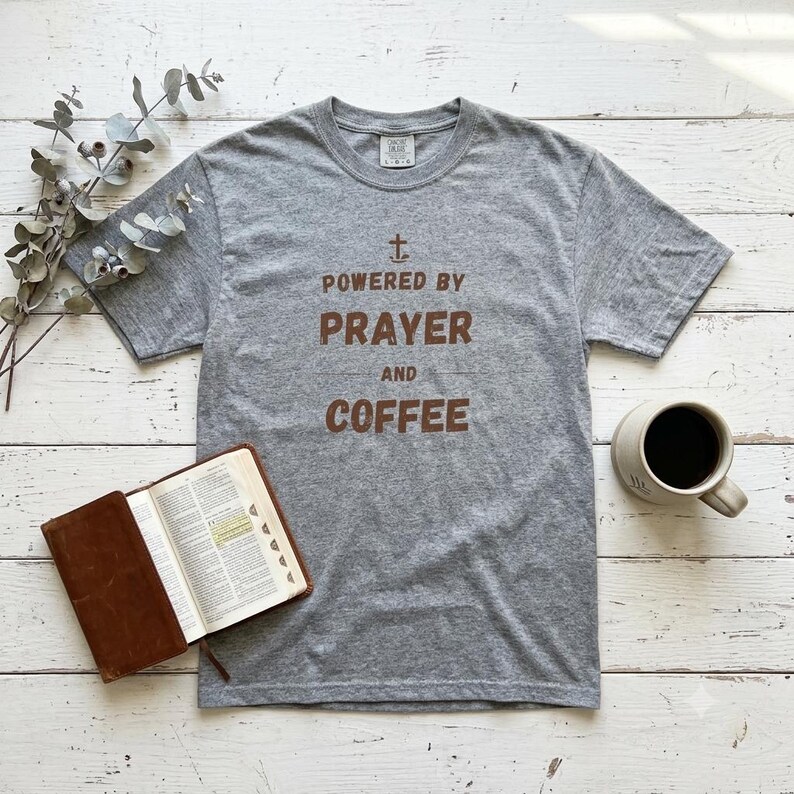 May include: A heather gray short-sleeve t-shirt featuring a brown graphic print that reads "POWERED BY PRAYER AND COFFEE" with a small cross symbol above the text. The shirt has a crew neckline and a standard fit, displayed on a flat surface.