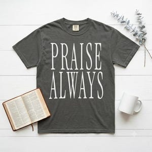 May include: A charcoal gray short-sleeve t-shirt featuring the white text PRAISE ALWAYS printed in a tall, serif font on the chest. The shirt has a crew neckline and a standard fit, displayed flat with a book and a white mug nearby.