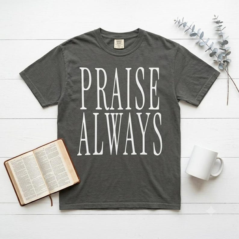 May include: A charcoal gray short-sleeve t-shirt featuring the white text PRAISE ALWAYS printed in a tall, serif font on the chest. The shirt has a crew neckline and a standard fit, displayed flat with a book and a white mug nearby.