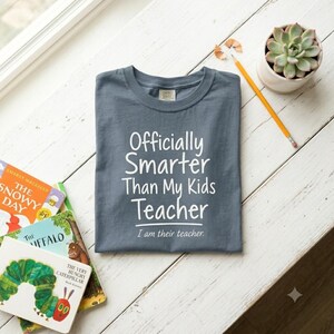 May include: A slate blue crew neck t-shirt featuring the white printed text "Officially Smarter Than My Kids Teacher" followed by the smaller text "I am their teacher." The shirt is displayed alongside a yellow pencil and a small succulent plant.