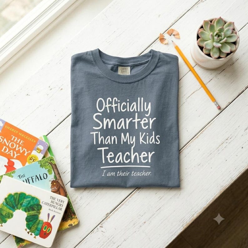 May include: A slate blue crew neck t-shirt featuring the white printed text "Officially Smarter Than My Kids Teacher" followed by the smaller text "I am their teacher." The shirt is displayed alongside a yellow pencil and a small succulent plant.