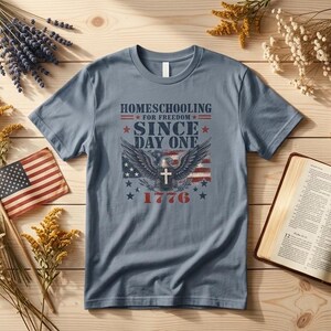 May include: A slate blue short-sleeve t-shirt featuring a graphic print that reads "HOMESCHOOLING FOR FREEDOM SINCE DAY ONE 1776" with an eagle, a cross, and American flag elements. The shirt is displayed flat with a small flag and an open book.